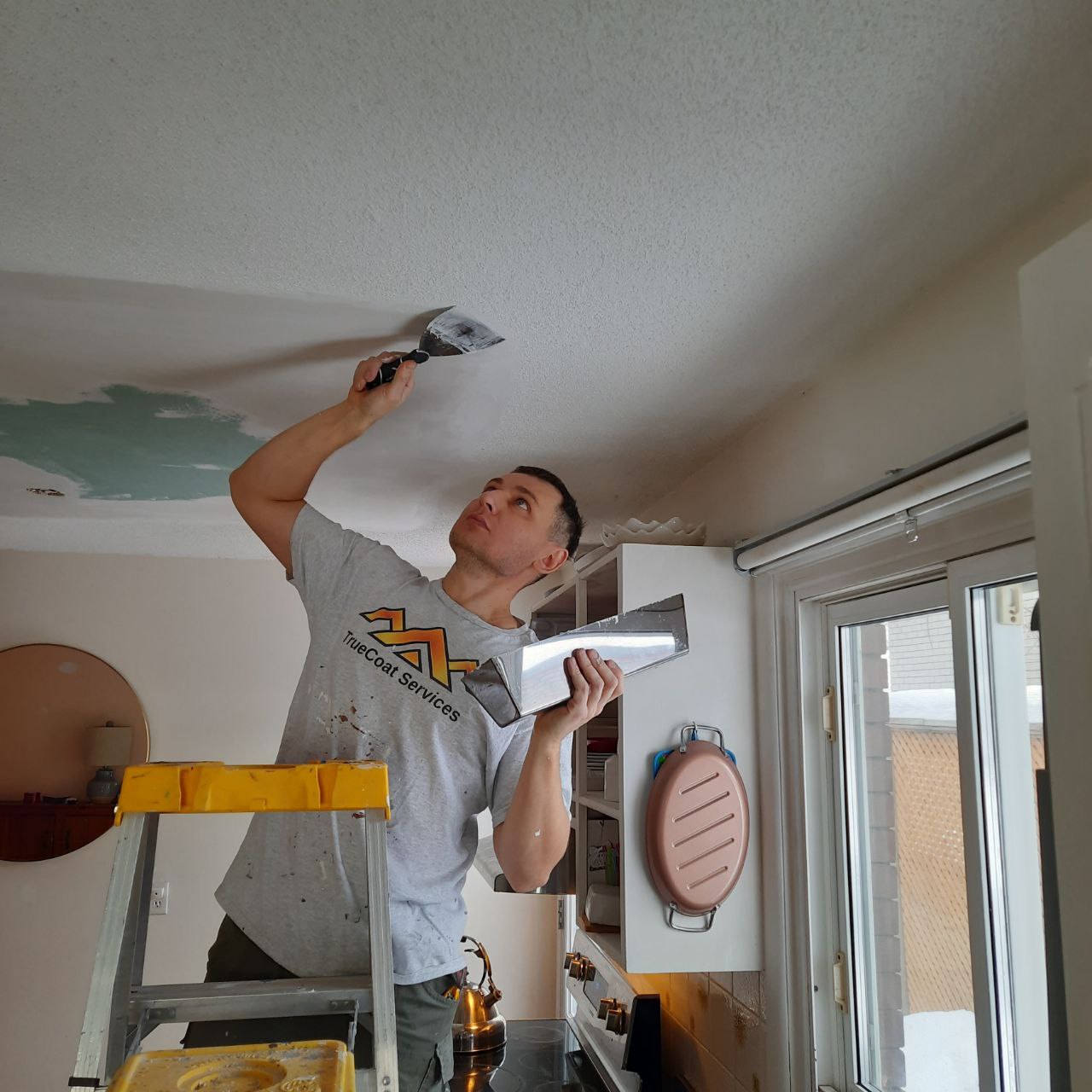 TrueCoat tradesperson skim-coating a repaired ceiling with a wide taping knife before repainting